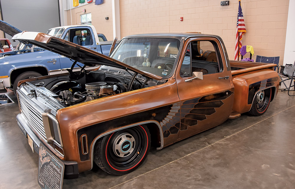 A highly customized, lowered copper-patina Chevrolet pickup truck with black wing graphics and the hood open, displayed indoors.