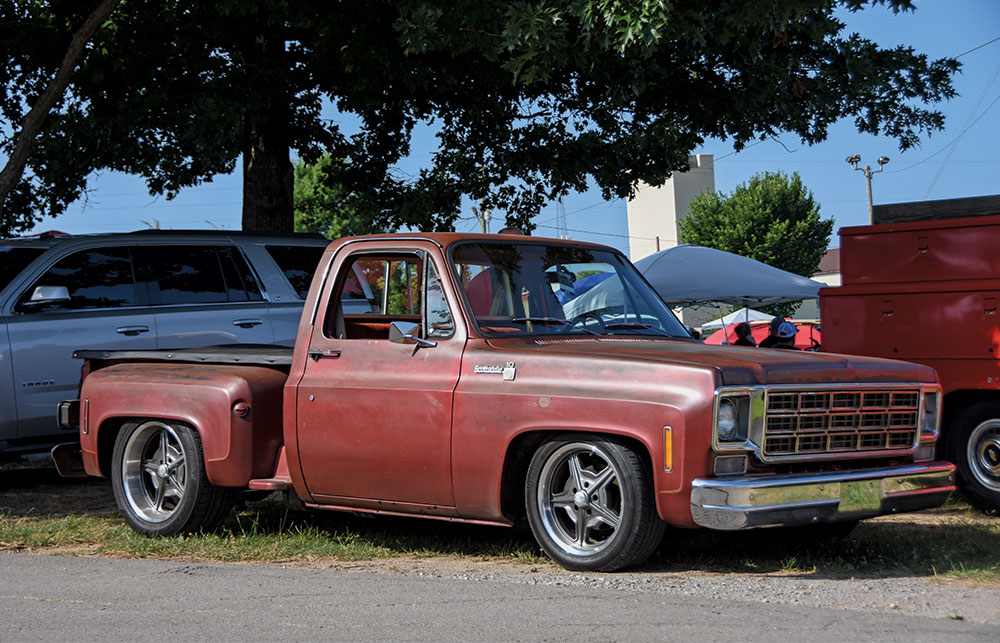 A custom, lowered red patina Chevrolet C10 pickup truck with large, stylized five-spoke wheels, parked outdoors under a tree.