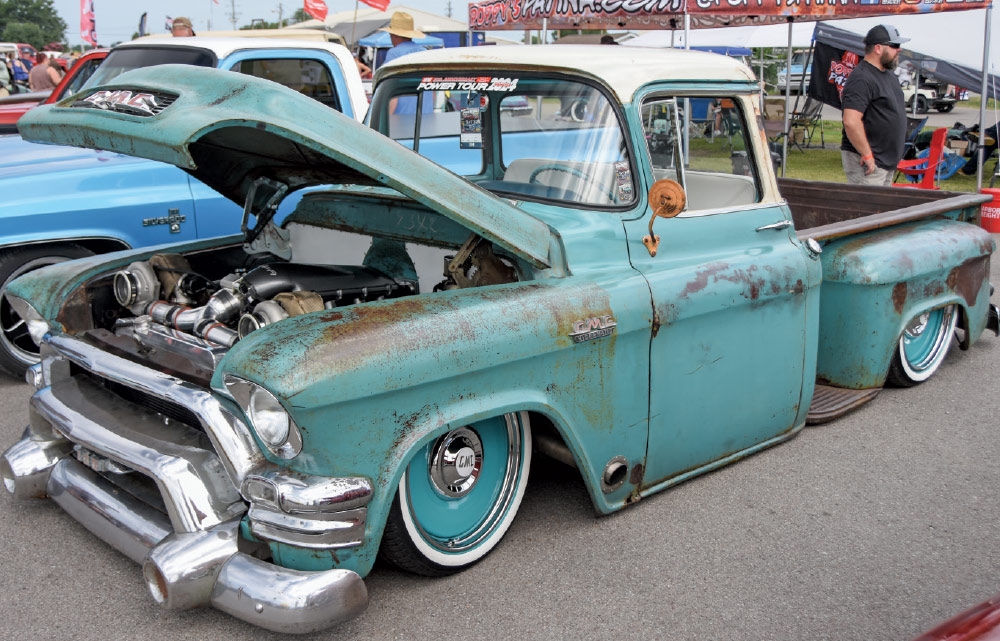 A lowered, patina turquoise GMC pickup truck with its hood open, revealing a custom engine, and painted wheels at a car show.