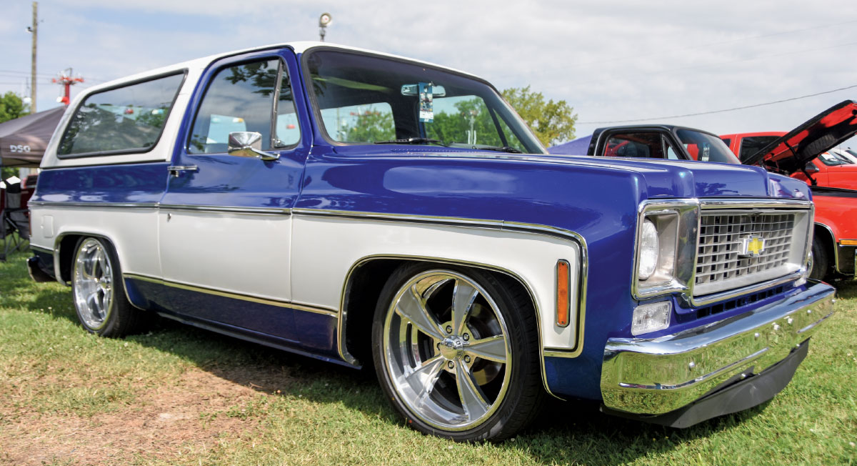 A classic, low-riding blue and white Chevrolet K5 Blazer with chrome five-spoke wheels, parked on the grass at an outdoor event.