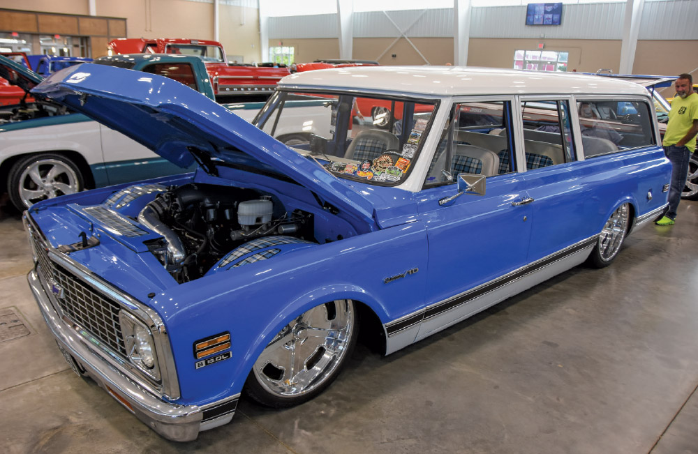 A highly customized, lowered blue and white Chevrolet Suburban with the hood open, massive chrome wheels, and tartan plaid interior accents.