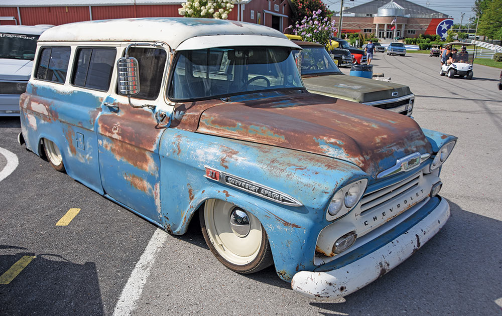 A slammed, patina blue and rust Chevrolet Apache Suburban with white-painted wheels, parked at an outdoor car show.