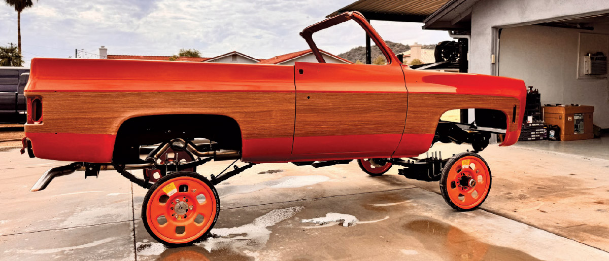 A stripped Chevrolet C10 body shell painted orange and wood-grain, sitting on a custom chassis with orange wheels, in a driveway.