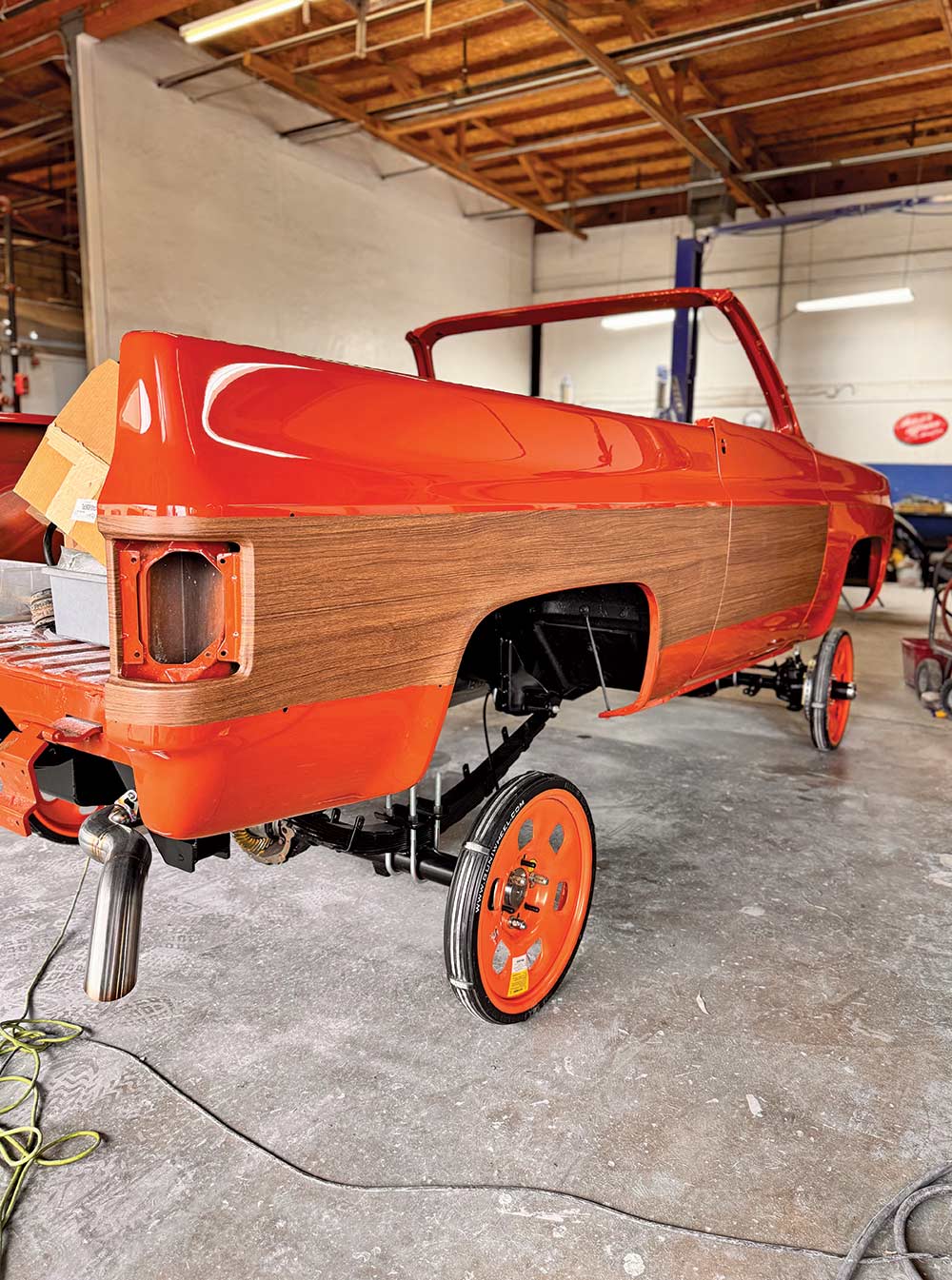The rear view of an orange Chevrolet C10 body shell with wood-grain side paneling, mounted on a chassis with bright orange wheels in a shop.