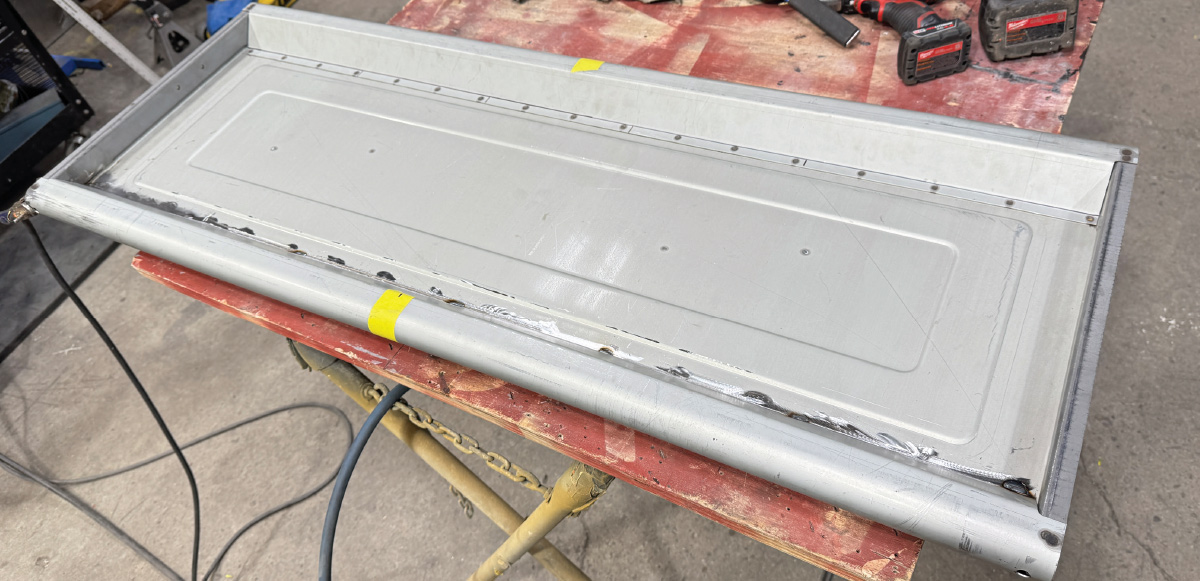 Tailgate on reddish worktable, perimeter weld seams visible, tools scattered, concrete floor, vehicle tires behind