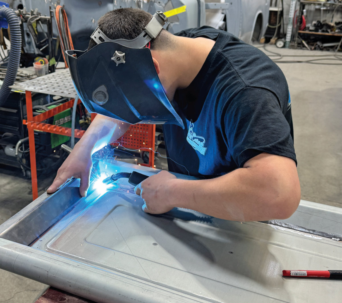 MIG welding inner edge, helmet on, clamps and marker on table, shop racks