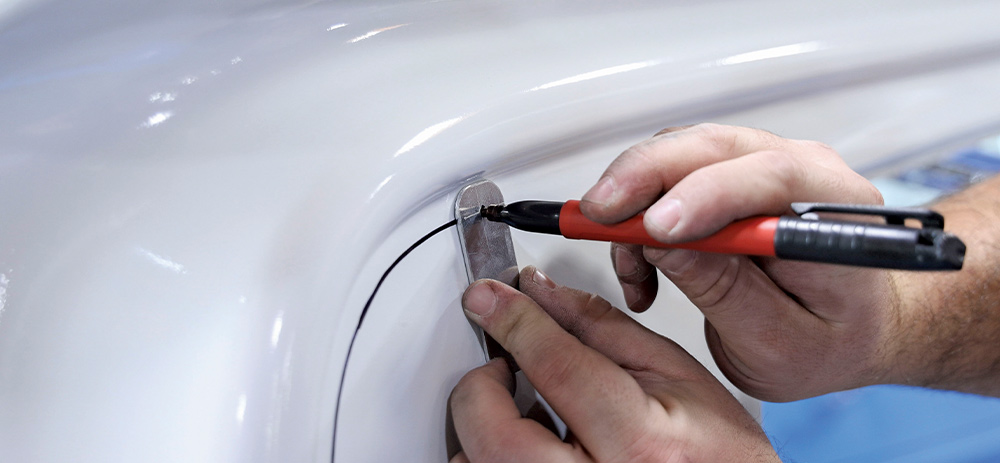 A close-up of a person's hands using a red marker and a small metal template to trace a curved line onto a freshly painted white vehicle body panel, likely marking a cut-out area.