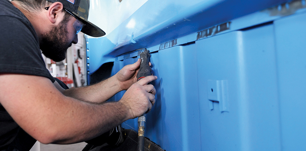 A man with a beard and baseball cap uses an air-powered oscillating tool to grind a seam on the light blue painted exterior body of a classic pickup truck in a shop environment.