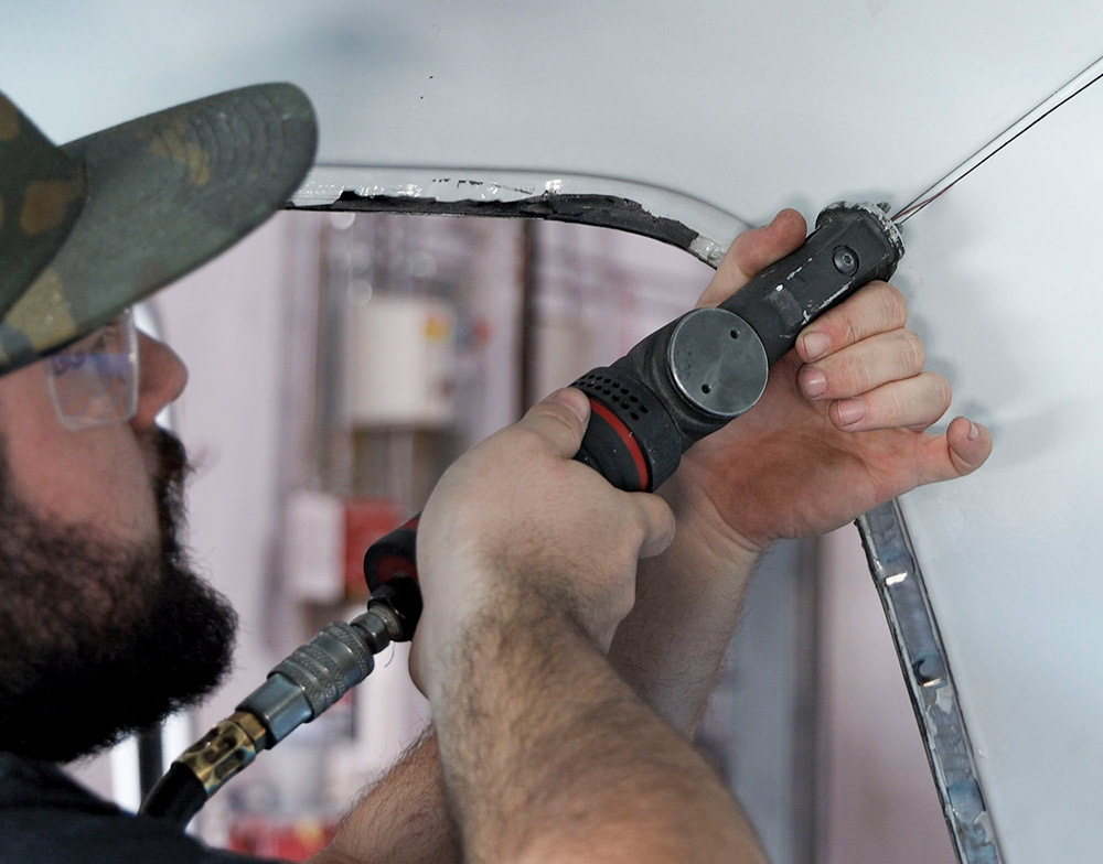 A man in a camouflage hat uses an air-powered oscillating grinding tool to carefully work on the painted blue metal in the door jamb area of a classic pickup truck.