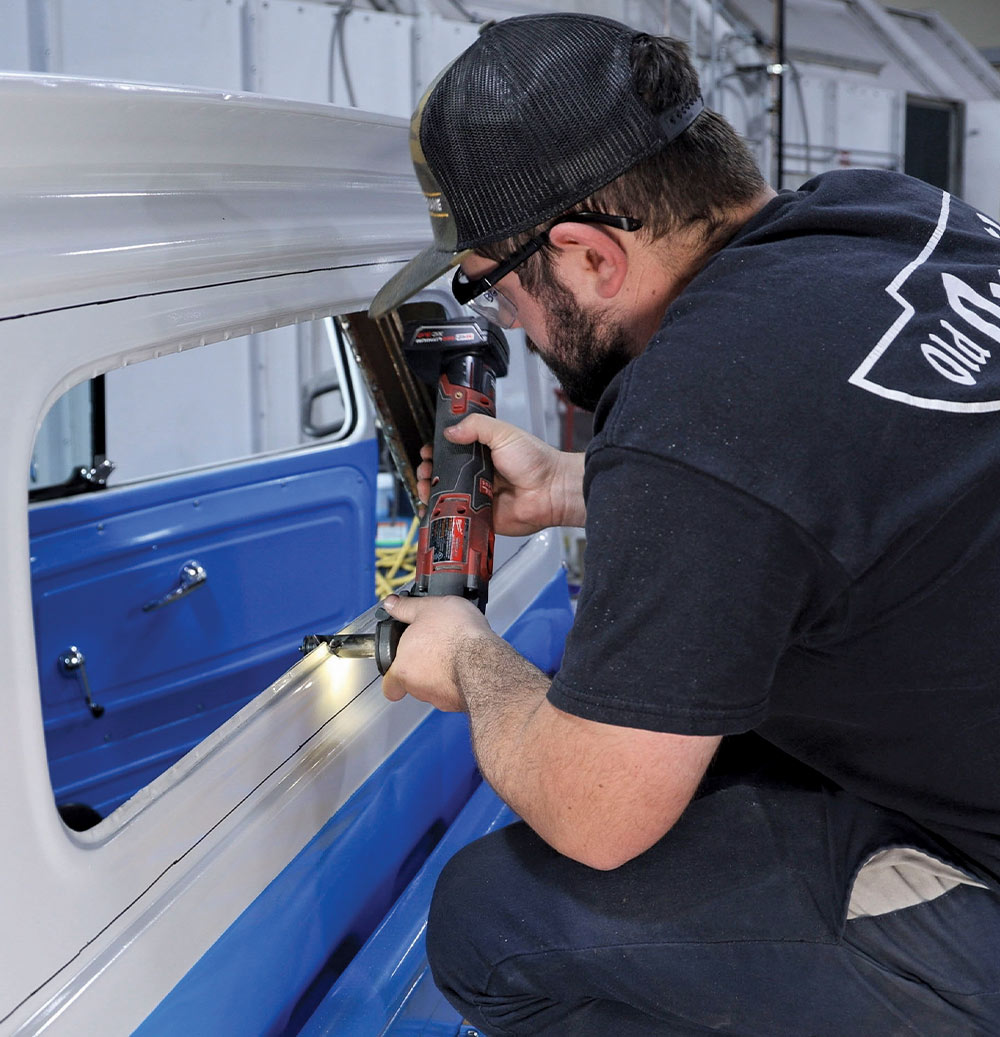 A man in a camouflage hat and black shirt uses a yellow-handled flat tool to scrape off old sealant and rust from the curved metal flange of a classic blue and white pickup truck's cab behind the driver-side door.