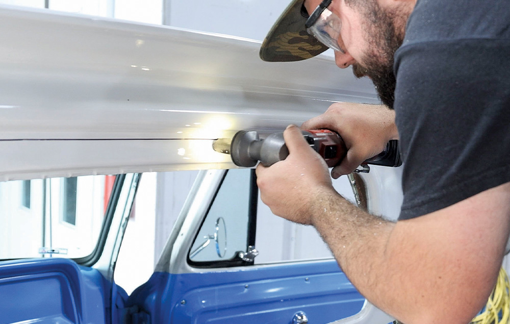 A man in a camouflage hat and black shirt uses a red and black power tool with a cutting blade to trim the seam around the lower edge of the rear window opening on a blue and white classic pickup truck cab.