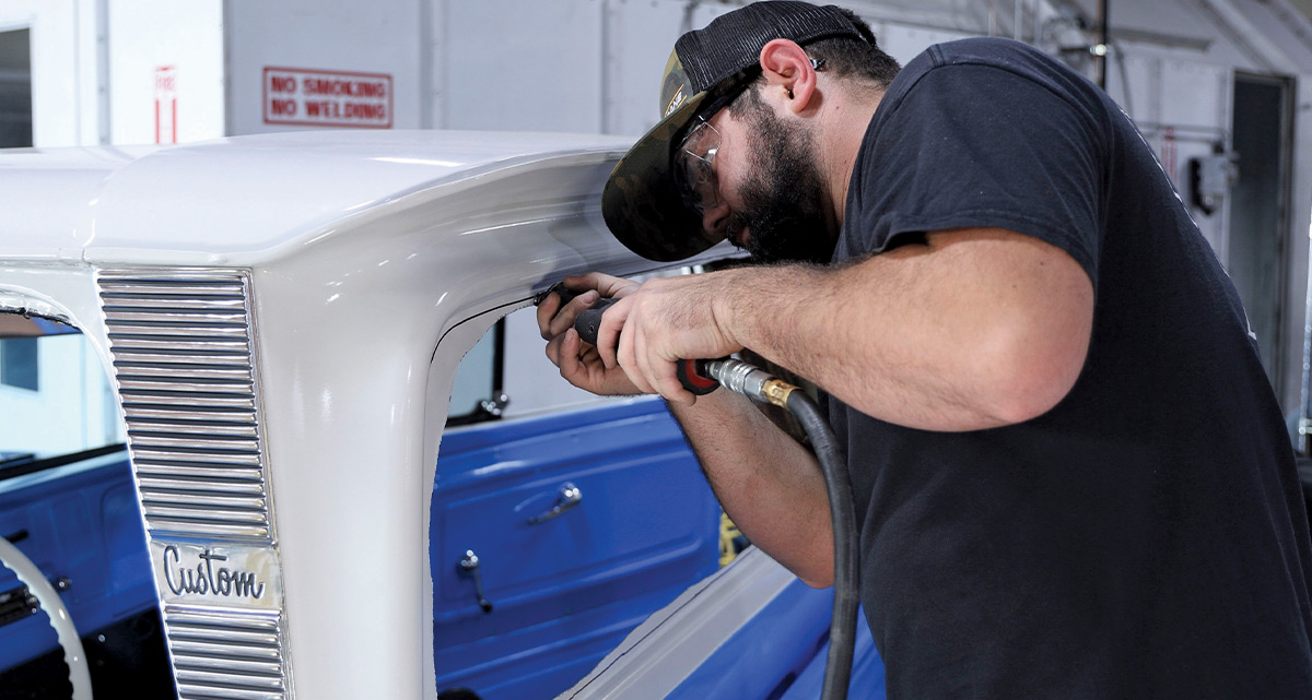 Close-up view of a man using an air-powered hole saw to drill a circular hole into the painted white inner roof panel of a classic pickup truck's cab, above the windshield opening.