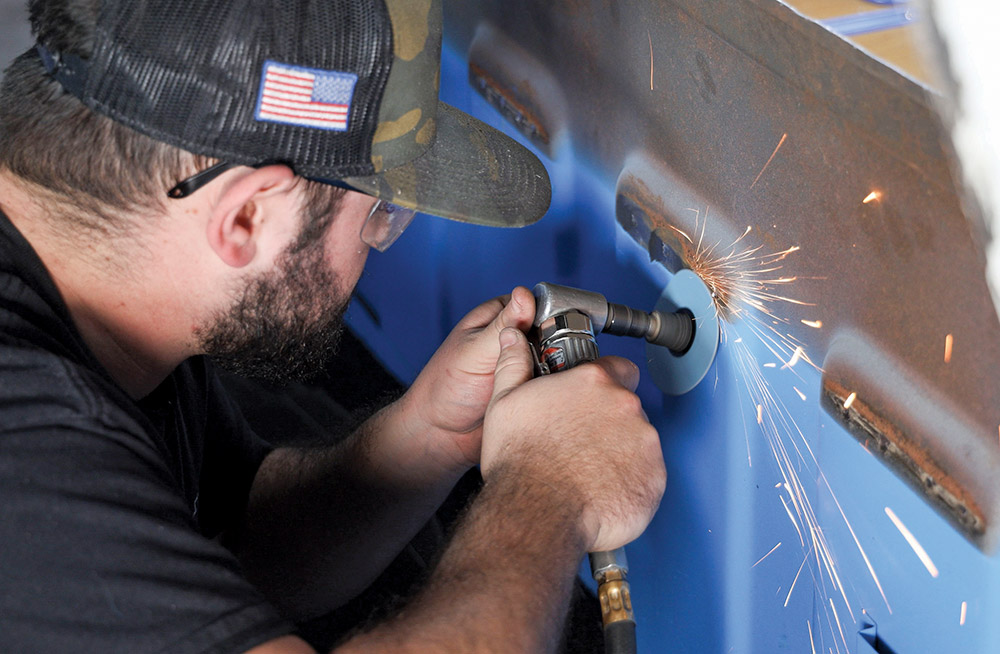 Close-up of a man using a high-speed rotary grinding tool to smooth a rough metal surface on a blue truck body, producing a shower of bright sparks from the friction.