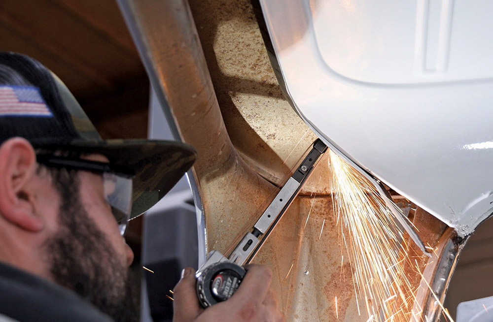 Close-up view of a man using a small rotary grinder on the rusted metal of a truck's door jamb near the hinge, creating a stream of bright sparks. He is wearing safety glasses and a camouflage hat.