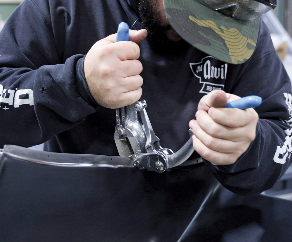 Close-up view of a man's hands using a large, specialty metal-bending tool to crimp the edge of a curved, glossy black spoiler or trim piece, likely preparing it for installation.