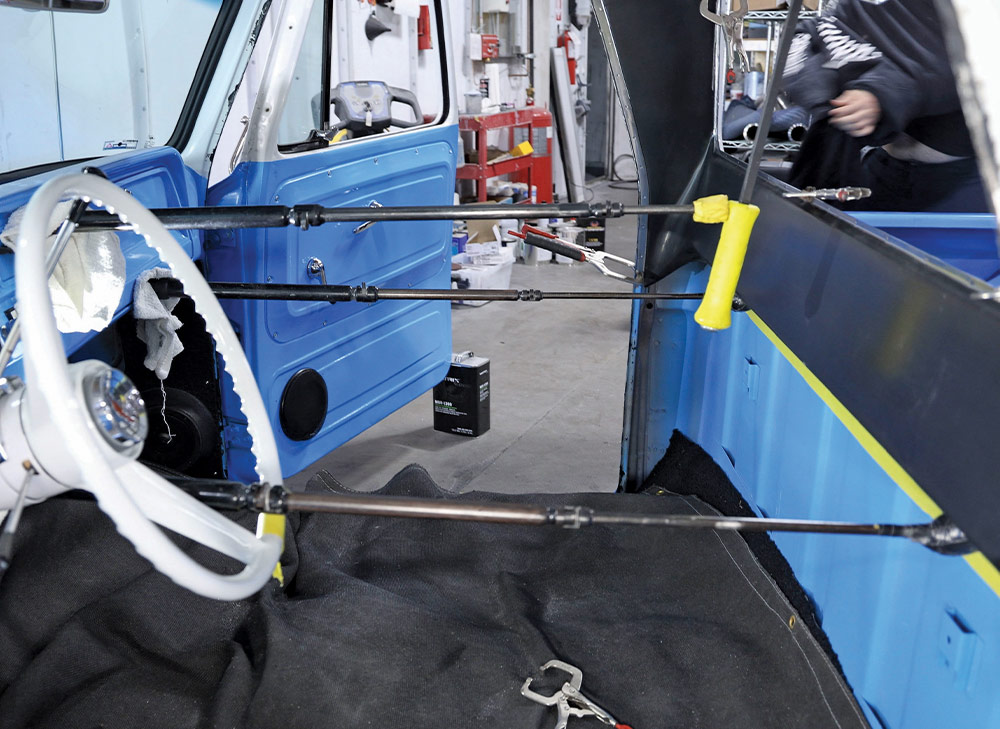 A wide shot of a truck interior showing the cab's bright blue side panels and door, with several horizontal rods temporarily holding the interior panels and a white steering wheel in the foreground.