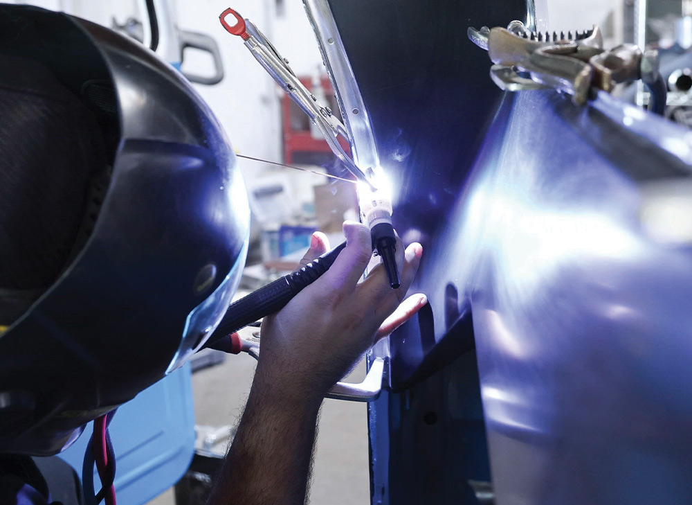 A close-up of a person wearing a welding helmet and gloves, TIG welding a seam on a metallic surface, with a bright arc and sparks visible from the torch tip.