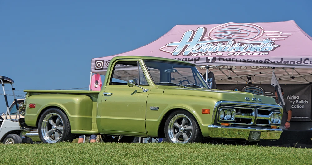Customized, glossy light green GMC pickup truck from the late 1960s or early 70s, featuring a lowered stance and custom gray five-spoke wheels, parked on grass beneath a "Harrison's Rod & Custom" tent at a truck fair.