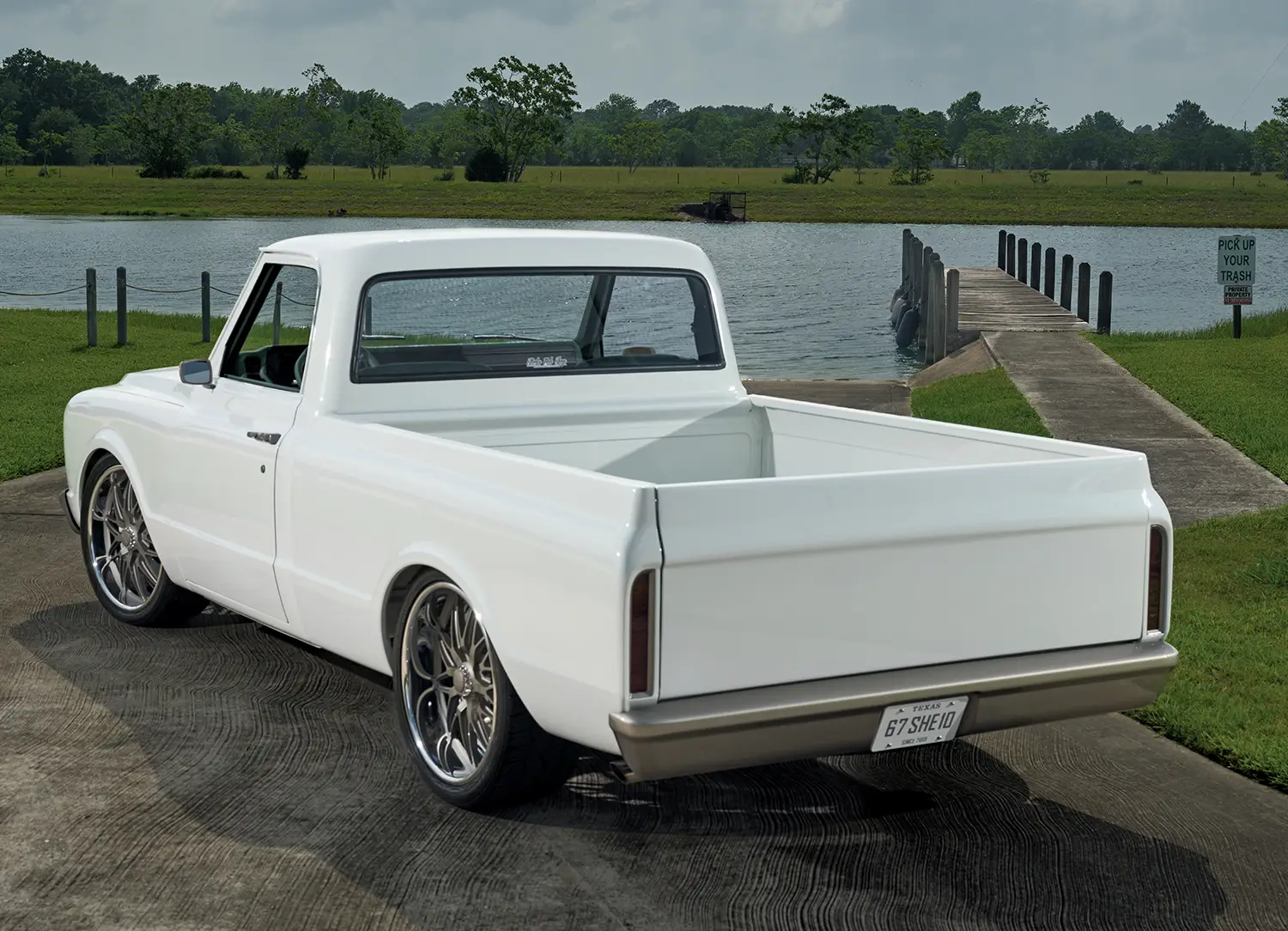 Rear three-quarter view of a customized, glossy white 1960s Chevrolet C-10 pickup truck with a lowered tailgate and bronze rear bumper, parked on a paved ramp overlooking a lake and pier.