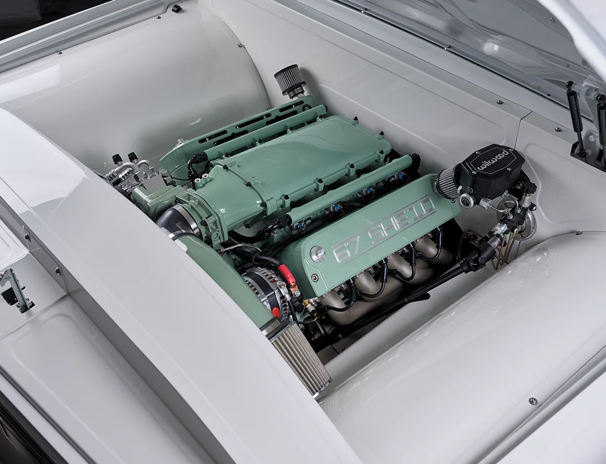 A detailed view of a classic truck's engine bay, featuring a custom light mint-green engine cover labeled "67 SHEBO" over a bright white, clean engine bay.