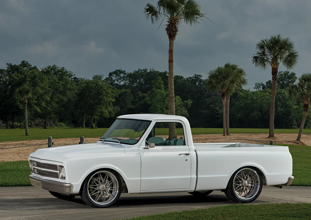 A fully restored, lowered white classic Chevrolet C10 pickup truck with large chrome aftermarket wheels, parked on a paved driveway with palm trees and a dark, dramatic sky overhead.