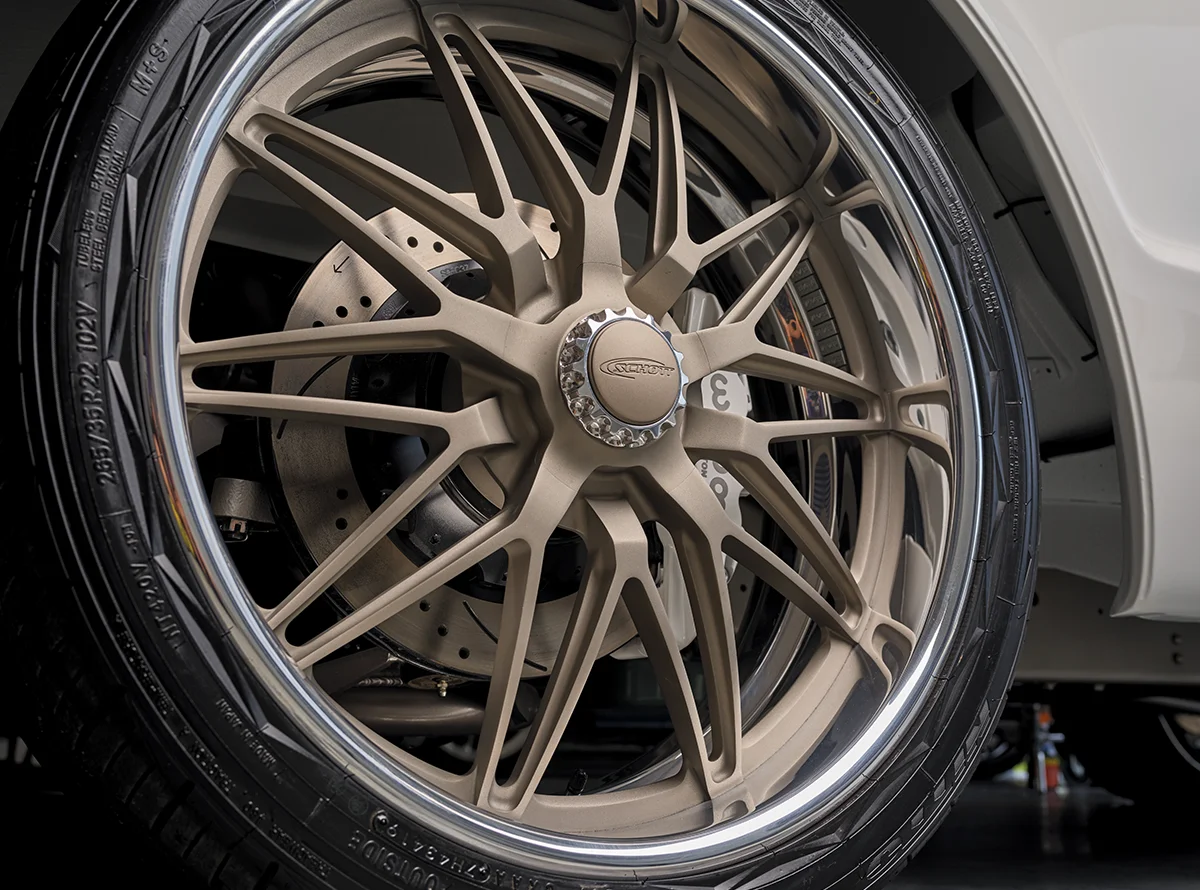 A close-up of a bronze-colored, multi-spoke aftermarket wheel and low-profile tire on a white vehicle, showing a large perforated brake rotor and caliper assembly visible behind the spokes.