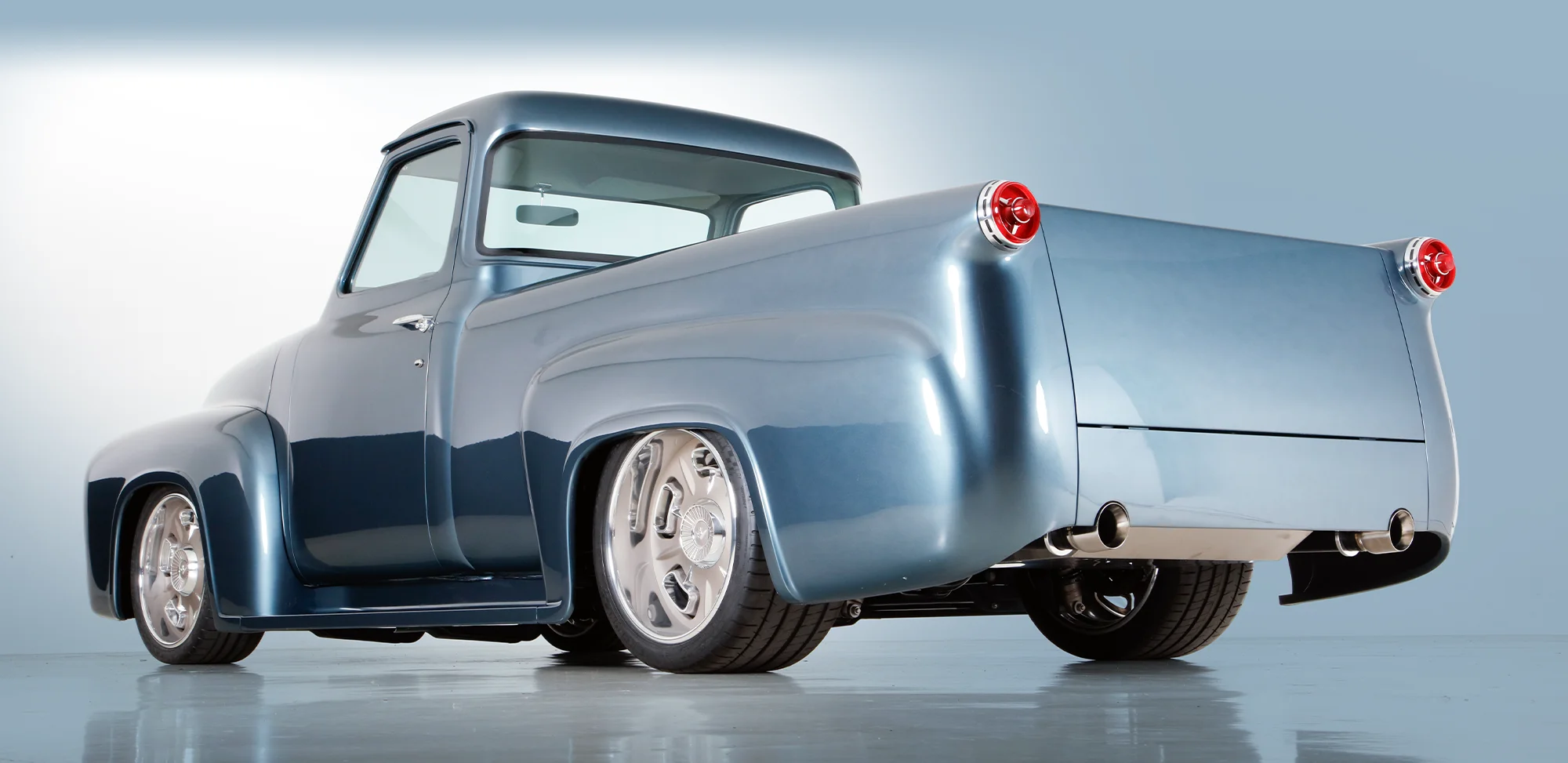 Rear three-quarter view of a metallic blue custom 1950s pickup truck with smoothed tailgate, integrated exhausts, and large polished wheels.