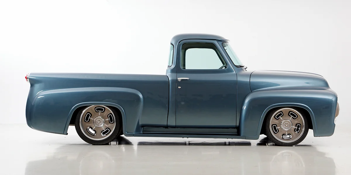 Side profile of a stunning metallic blue custom 1950s Ford F-100 style pickup truck with a lowered suspension and polished wheels.
