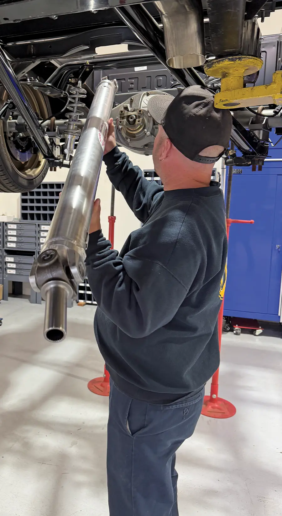 Mechanic installing a custom aluminum driveshaft underneath a classic truck on a lift, part of a high-performance drivetrain upgrade.