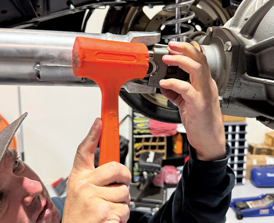 Mechanic using an orange rubber mallet to carefully tap the U-joint yoke of a custom driveshaft into place on the rear differential flange.