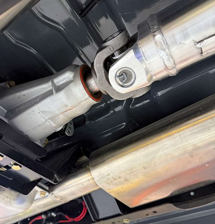 Close-up view of a new or custom driveshaft with a U-joint connecting to the rear of a transmission, positioned above a stainless steel exhaust system on a vehicle undercarriage