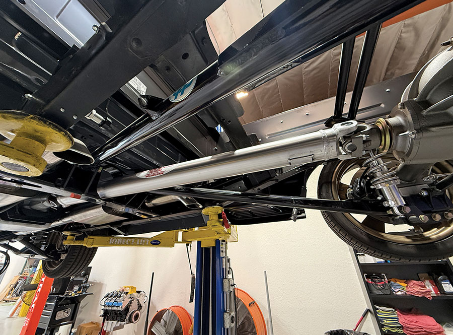 Full view of a custom aluminum driveshaft installed on a classic car or truck, connecting the transmission to the rear differential, seen from underneath the vehicle on a lift