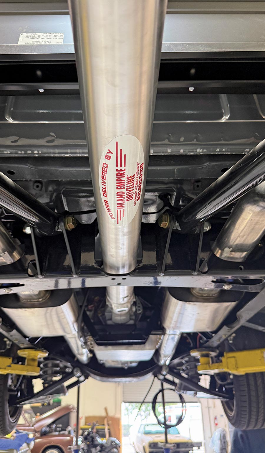 Undercarriage view of a classic or custom vehicle on a lift, showcasing a bright aluminum driveshaft, dual stainless steel exhaust system, and rear suspension components