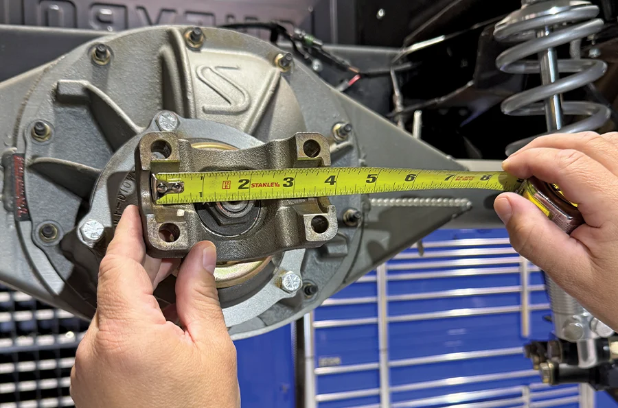 Hands using a measuring tape to measure the distance across a differential yoke on a Strange Engineering rear end, with coilover shocks visible in the background