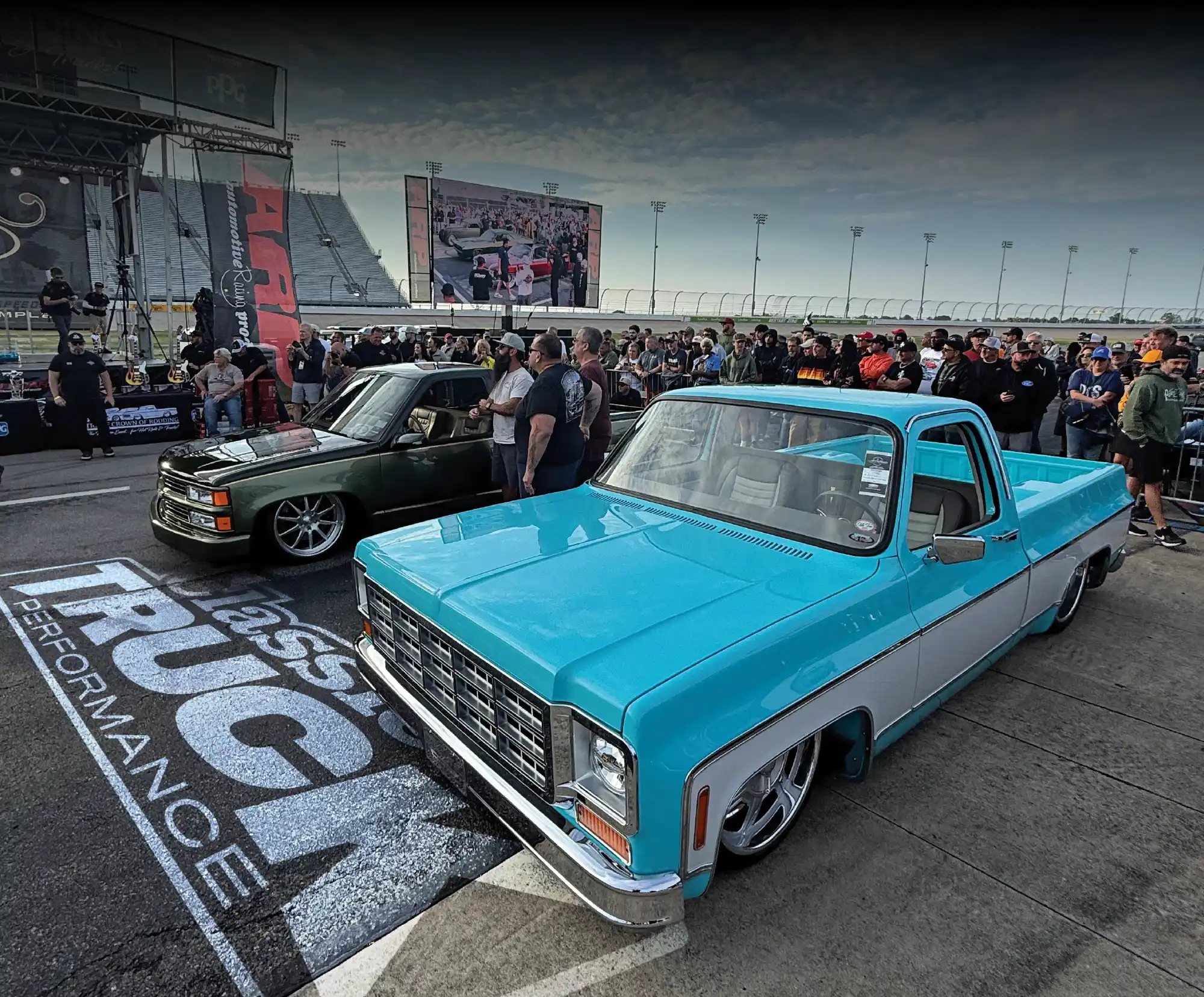 Two lowered classic pickup trucks, one blue/white and one green, are parked at a racetrack event with crowds and large screens in the background.