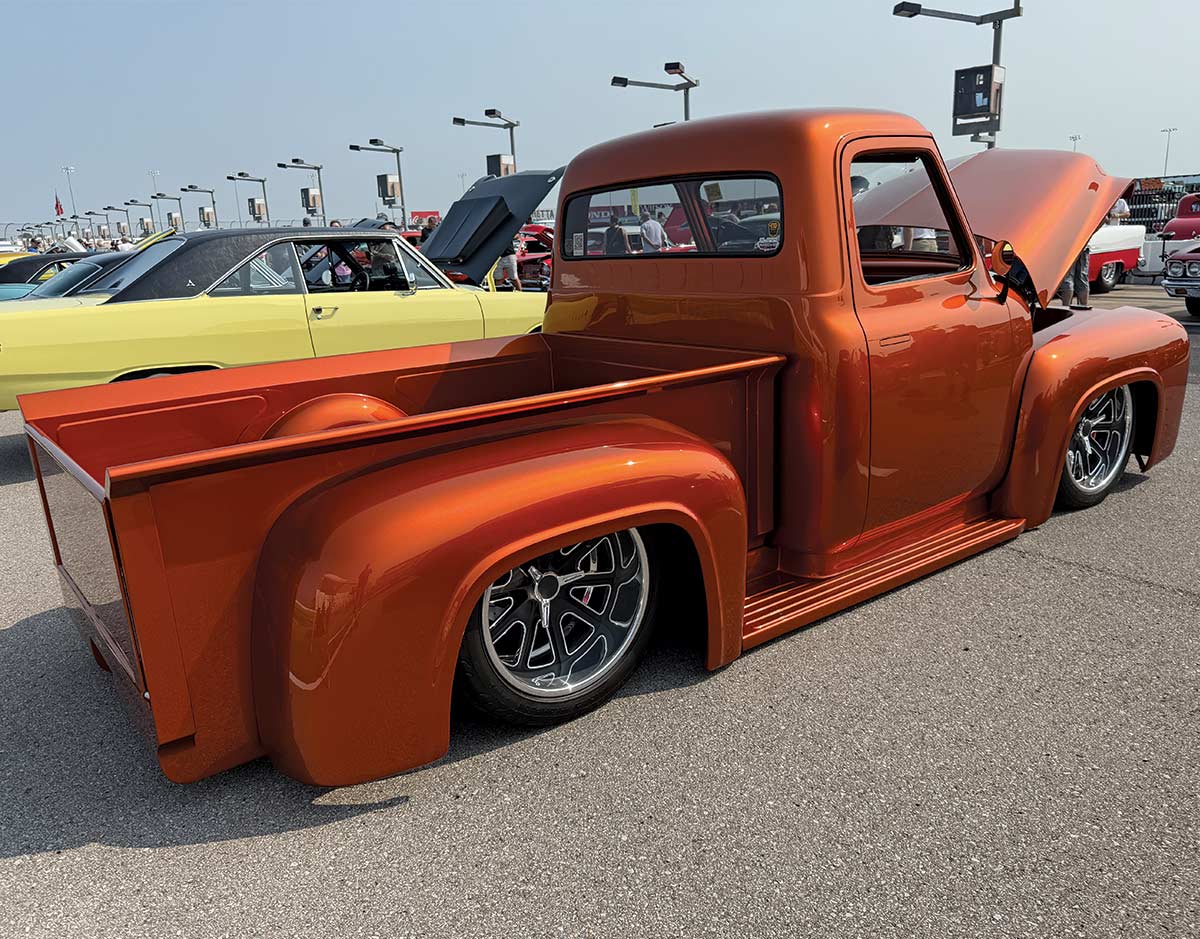 A custom, low-riding, metallic orange classic Ford F-100 pickup truck with the bed shaved and the hood open, on display at an event.