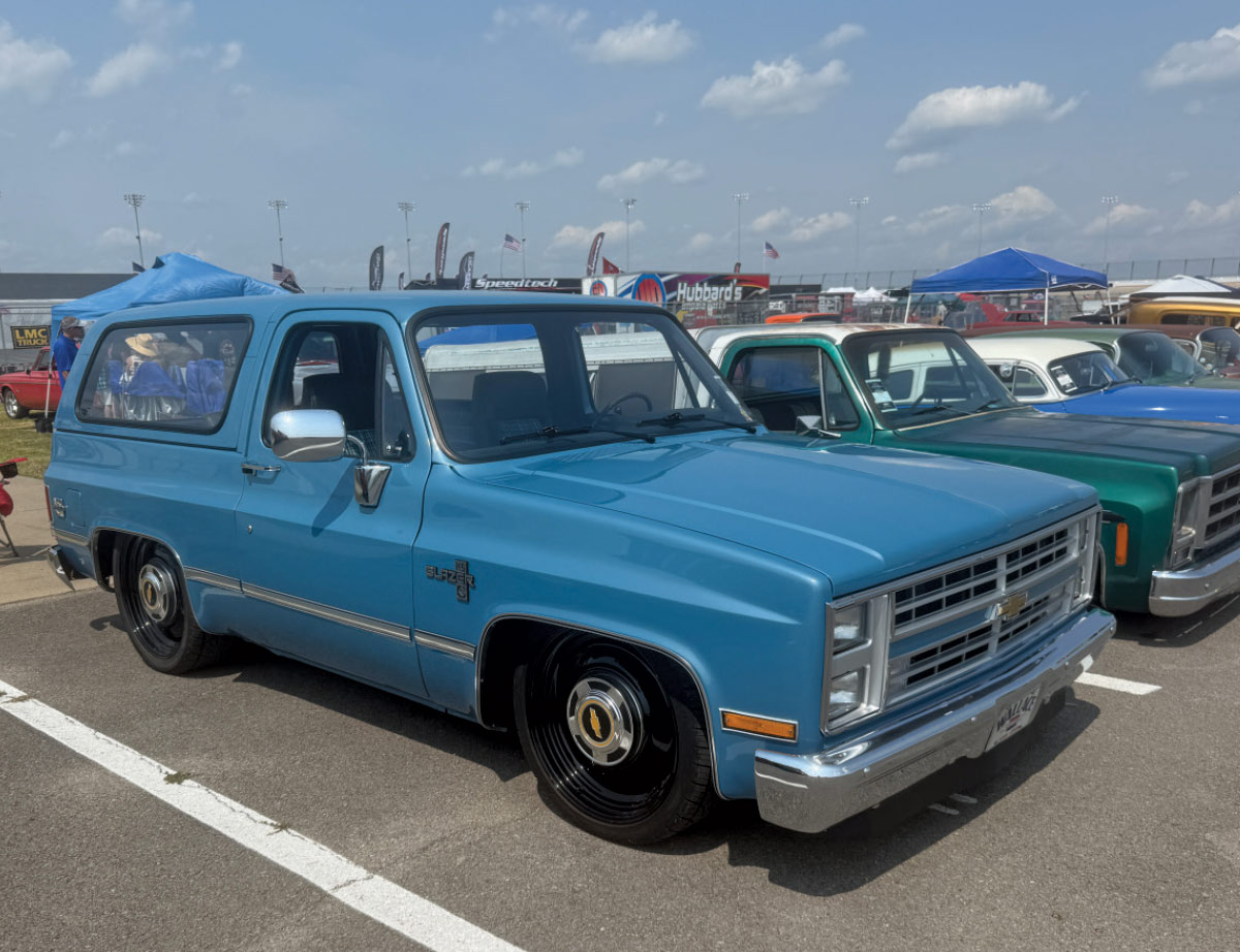 A lowered, light blue car with black wheels parked among other trucks at an outdoor event.