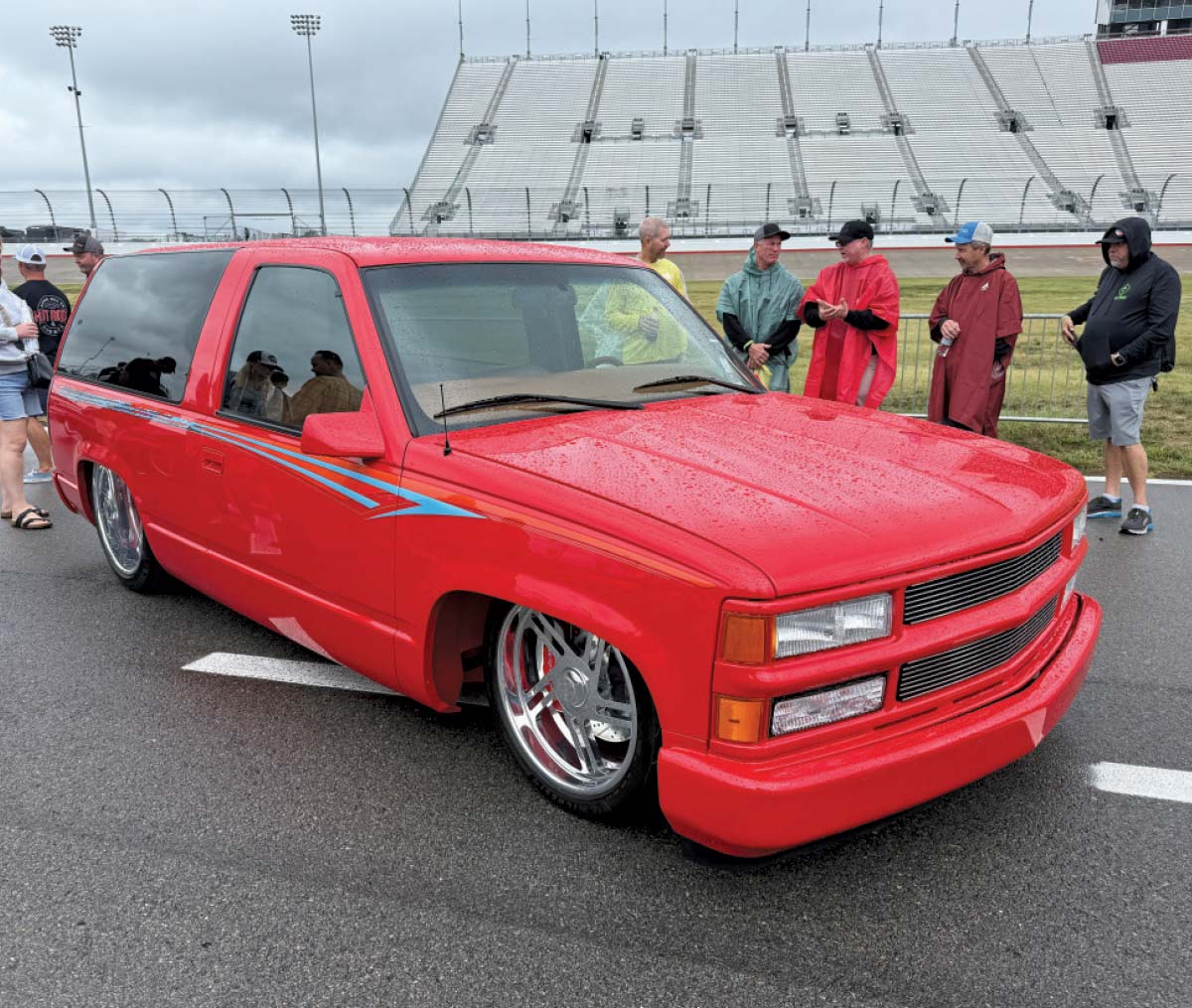  A heavily modified red K5 Blazer with pinstriping, a lowered suspension, and large chrome wheels is parked on a racetrack paddock.
