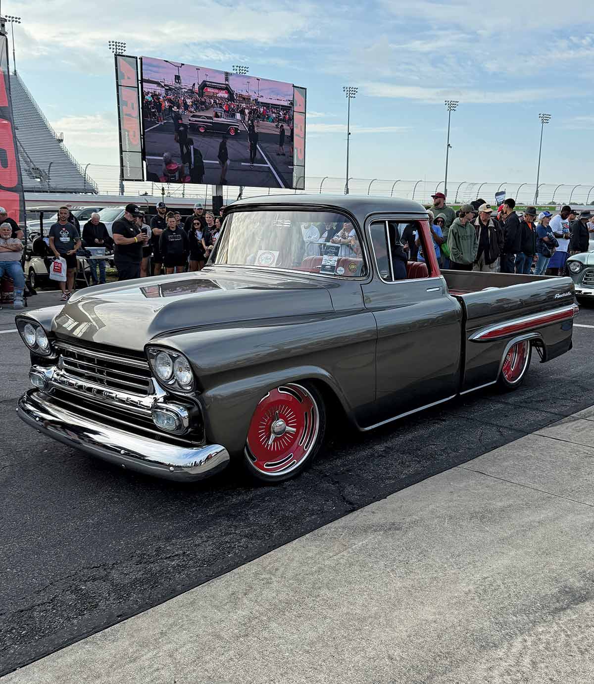A gray, lowered 1958 Chevrolet Apache pickup truck with red wheels and trim in front of a jumbotron.