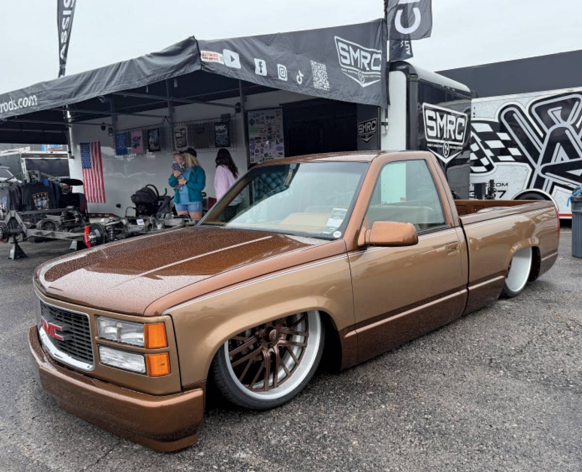 A lowered, two-tone brown GMC pickup truck with custom bronze wheels parked near a vendor trailer.