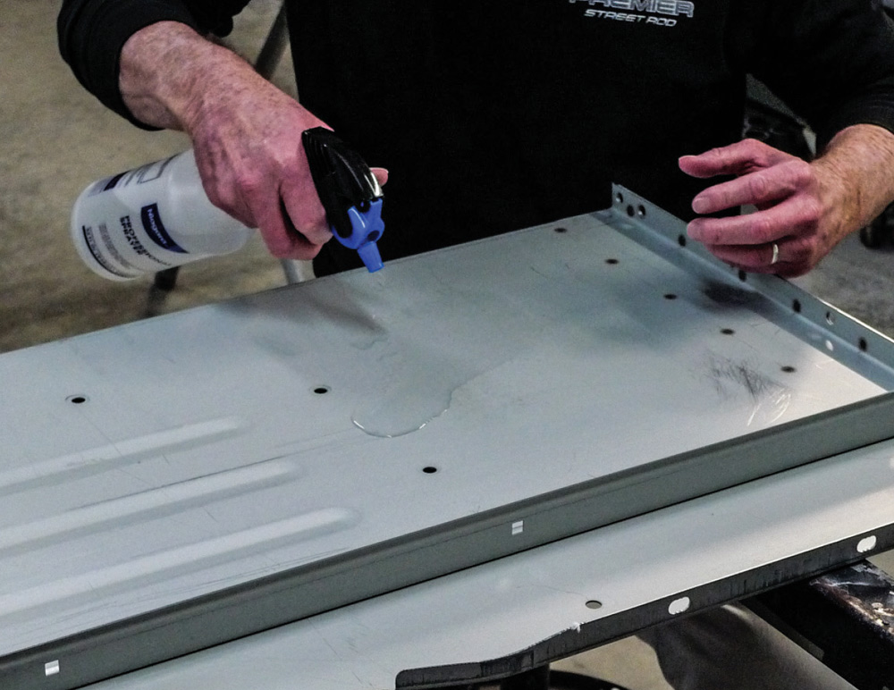 Technician sprays water onto a gray metal truck bed panel to test for cleanliness. The water sheets on the surface instead of beading up, confirming that all grease and wax residue has been successfully removed.