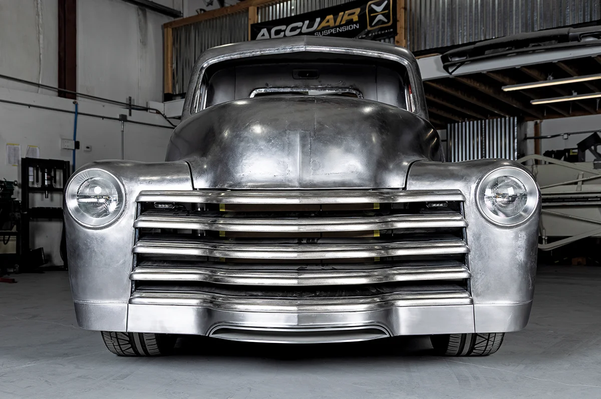 A vintage 1950s truck undergoing a metal-shaping restoration, showing the raw steel finish and headlights in a shop with an AccuAir Suspension banner