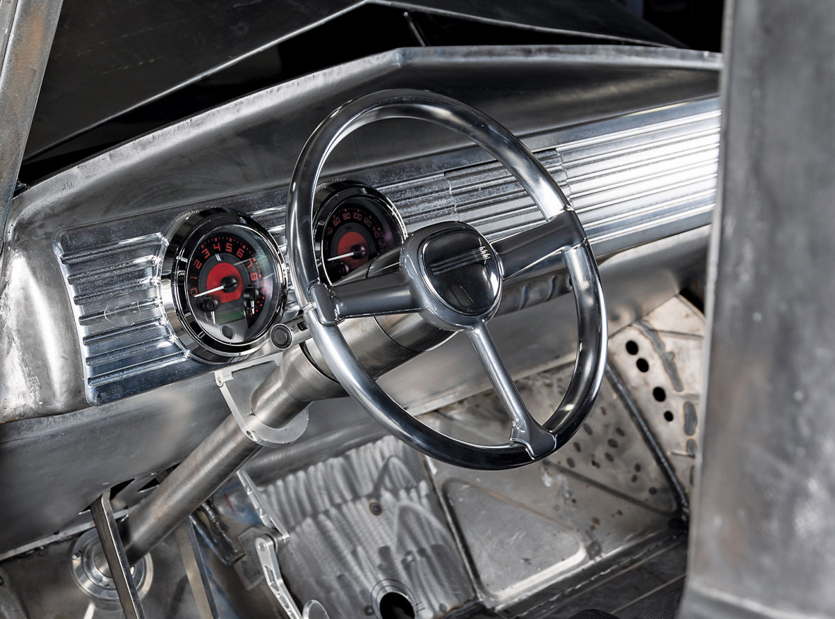 Interior of a bare metal truck showing a polished three-spoke steering wheel and two round gauges with red faces set into a metallic dashboard.