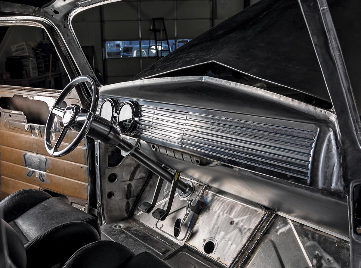 Wide interior view of a bare metal truck cab featuring black bucket seats, a chrome steering wheel, and exposed metal floor panels and pedals.