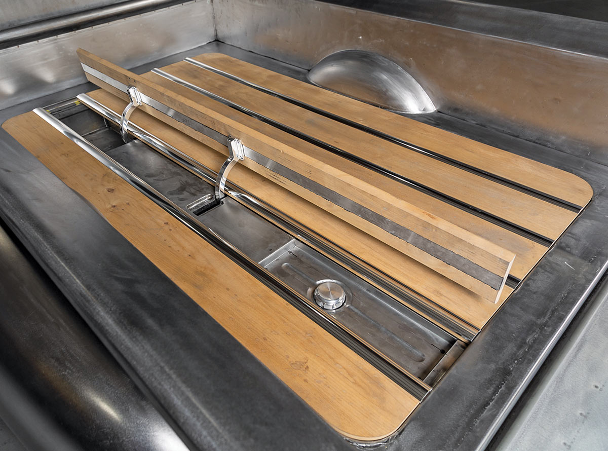 Close-up of a bare-metal truck bed featuring wood floor planks and polished metal divider strips. A center section of the wood floor is hinged open, revealing a recessed metal fuel filler cap in the bed floor.