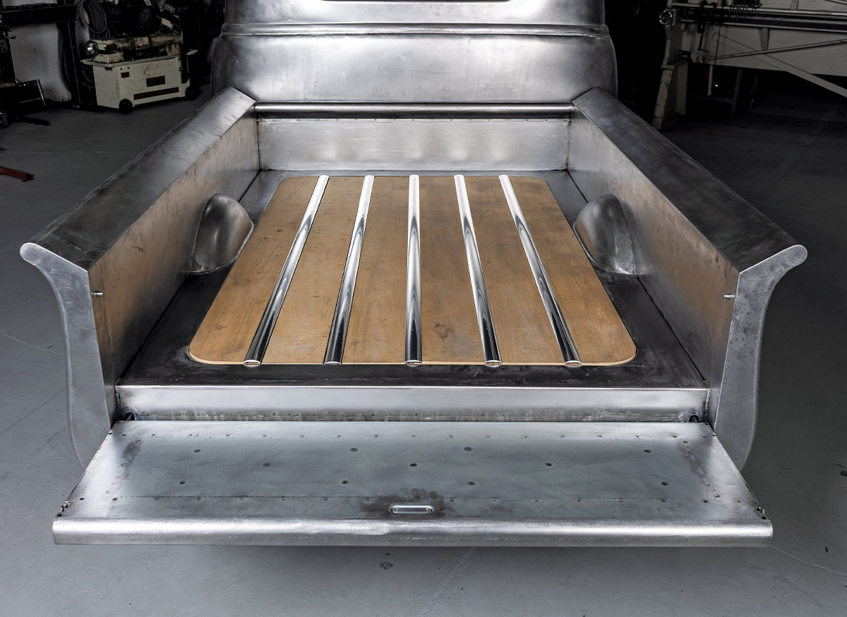 View of a bare metal truck bed with the tailgate lowered horizontally. The bed floor features wood planks and polished metal divider strips. The interior metal walls are unpainted with visible raw textures and weld marks.