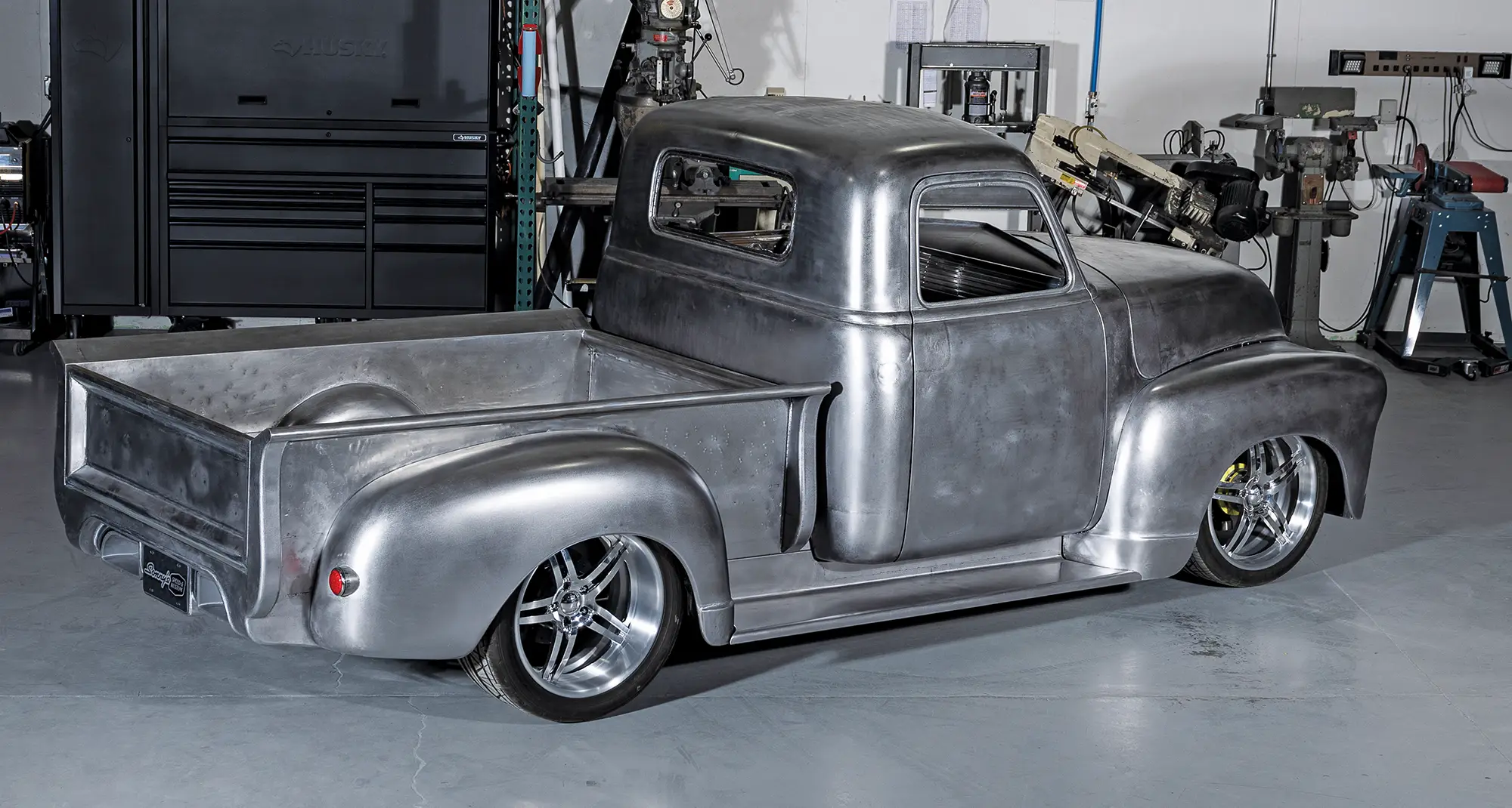 High-angle, rear three-quarter view of a bare-metal Chevrolet 3100 truck in a workshop. The truck features a lowered stance, chrome multi-spoke wheels, and raw metallic bodywork with visible sanding marks.