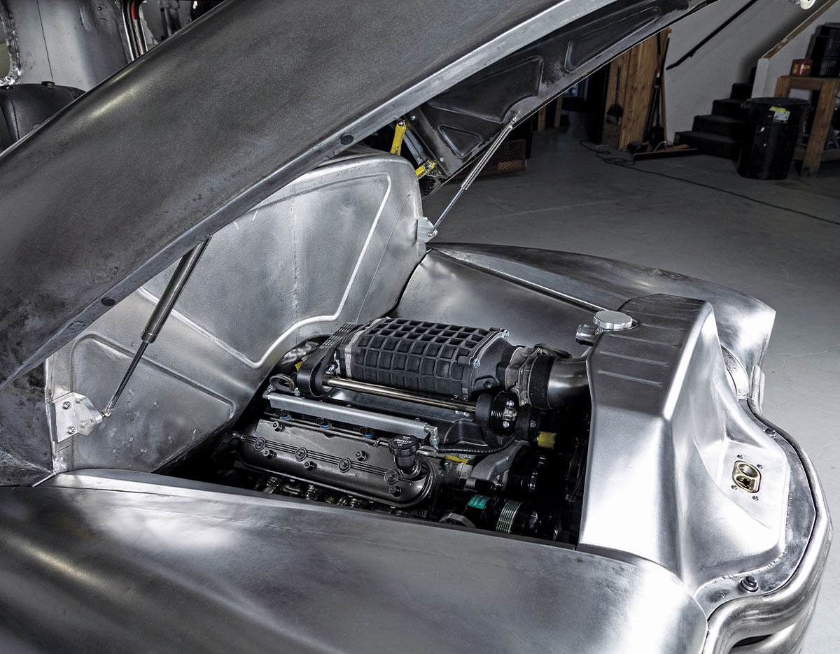 Engine compartment of a bare-metal truck with the hood held open by gas struts. The engine features a large black supercharger, surrounded by unpainted metallic inner fenders and a custom radiator shroud.