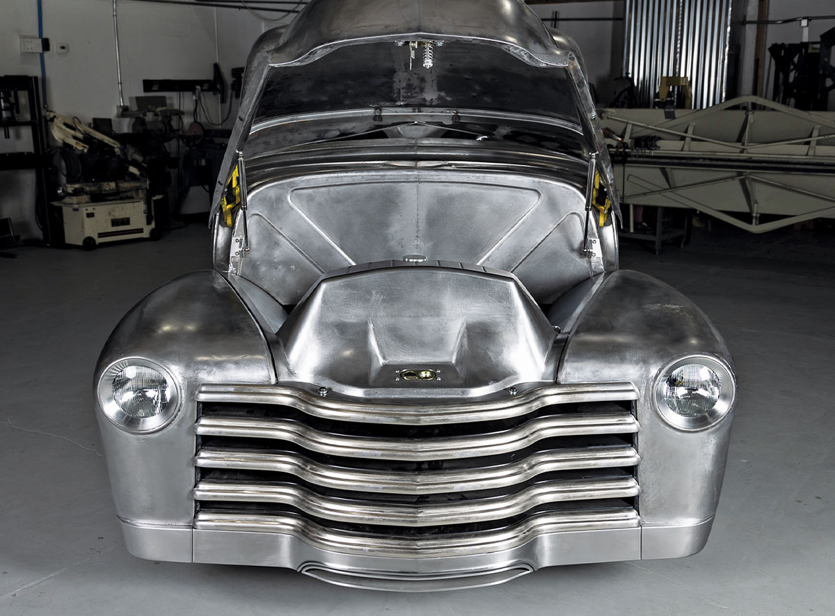 Front view of a bare-metal Chevy 3100 with the hood open. Inside the engine bay, a custom unpainted metal shroud covers the radiator area. The truck features a polished horizontal-bar grille and round headlights.