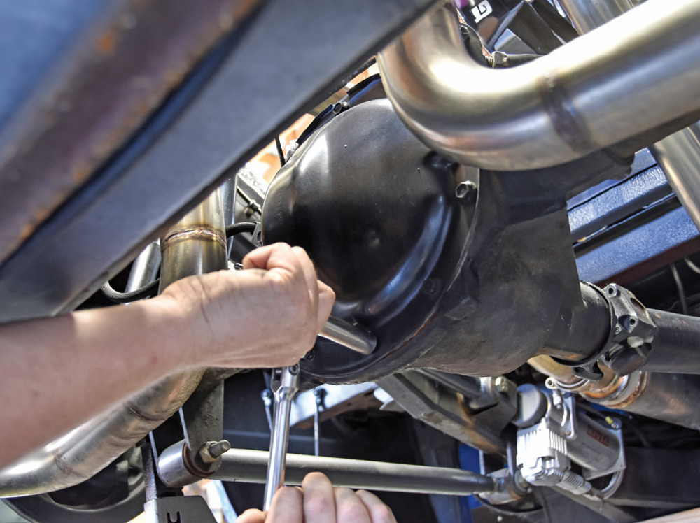 Hands using a socket wrench to unbolt a black differential cover on a vehicle's rear axle. Exhaust pipes and chassis components are visible under the blue vehicle body.